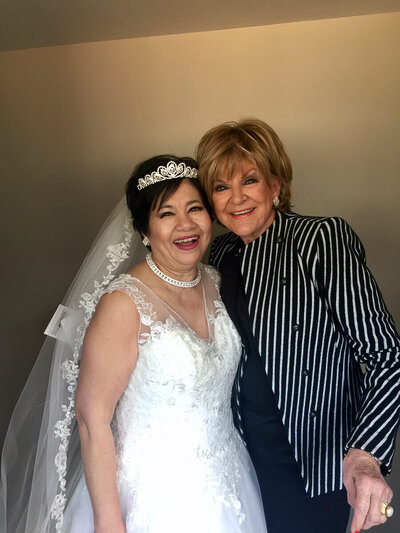 Author and speaker Nita Tin smiling broadly in a white lace wedding gown with tiara and pearls at her 50th wedding anniversary vow renewal, standing arm in arm with renowned Bible teacher Kay Arthur, who is smiling in a black skirt and a white-and-black dress blazer.