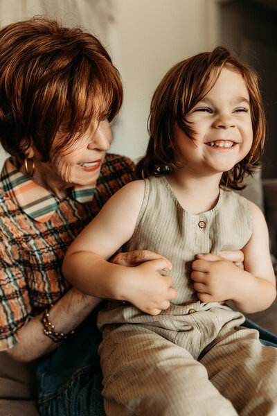A joyful toddler laughs while being held by their grandmother during a warm, light-filled in-home session in Scottsdale, Arizona. The image captures their playful bond and generational love in a cozy and candid moment.
