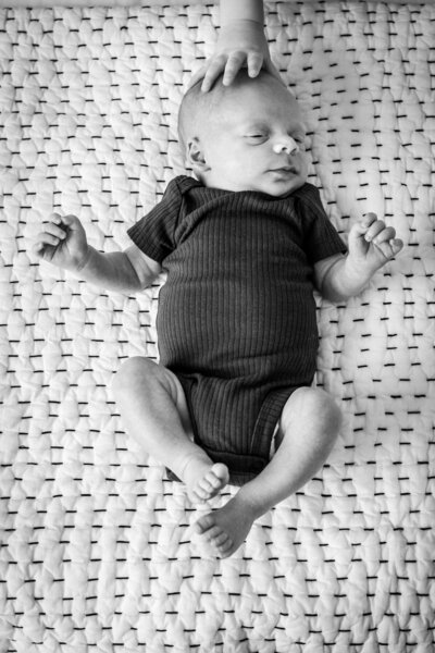 A black and white image of a newborn baby lying on a patterned blanket, with a small hand resting gently on the baby's head.