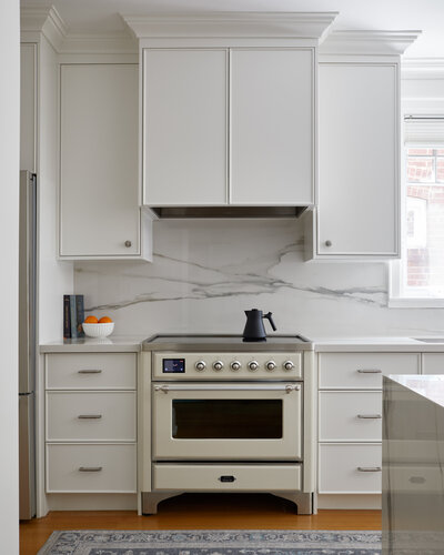 A bright airy kitchen with modern white cabinetry, wood flooring covered by a grey and white patterned rug, a luxurious marble backsplash, and white stove