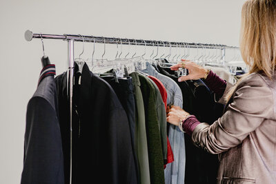 woman in a black suit sitting in front of a clothing rack filled with various outfits during a styling session