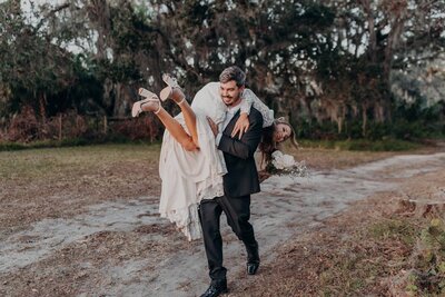 A groom carries a bride for a fun moment during sunset portraits at The Retreat, a Sarasota Wedding venue by preferred vendor, Angela Moon Photography. 