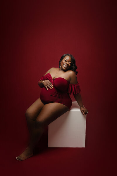 mom in red body suit on red backdrop for maternity session