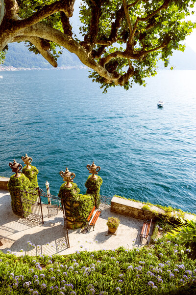 View of a serene lakeside with two ornate stone pillars covered in greenery, overlooking blue water. A small boat navigates the lake. Benches and vibrant plants surround the area, with tree branches extending above. Hills are visible in the distance.