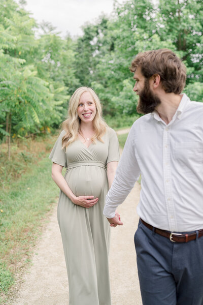 A father holds his pregnant wife's hand and leads her through green trees.