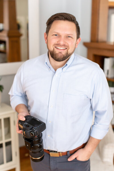 Brandon is smiling at the camera and is wearing a blue button down shirt and holding a camera