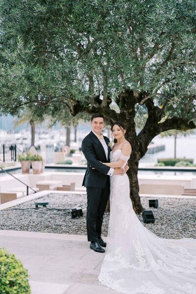 Bride and groom standing under olive tree with marina in the background, romantic photo by Toronto wedding photographer