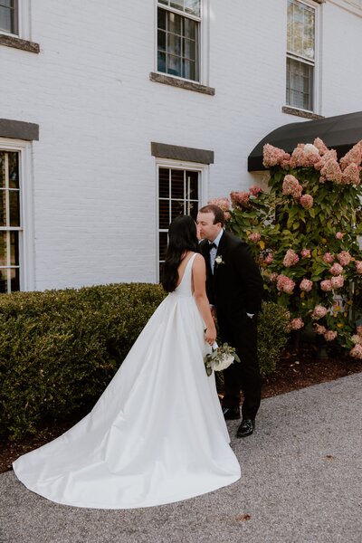 A bride and groom share a kiss outside the Stagecoach Inn, standing beside a bush of blooming hydrangeas. The bride wears a classic white gown with a deep V-back and a long train, while the groom is dressed in a black tuxedo. The couple is framed by the white brick exterior of the inn and the lush greenery, creating a romantic and timeless scene.