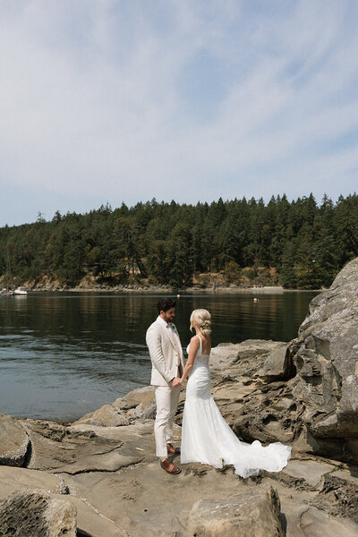 Bride and groom holding hands on rocks by the water.
