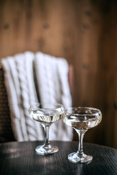 image of two champagne coupes on a black round table, with a chunky knit blanket over a brown wooden chair in the background