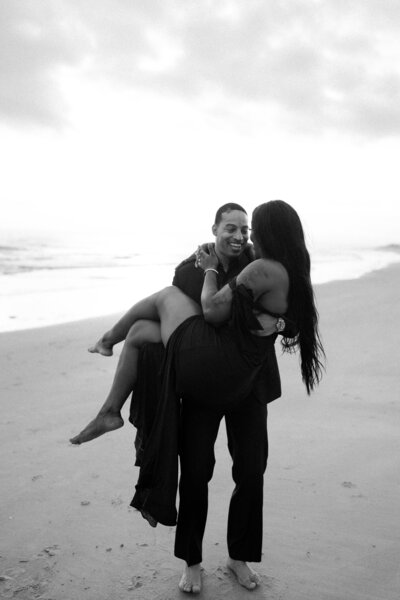 Black and white photo of a man carrying a woman on a beach, both smiling and wearing matching black outfits. Joyful and romantic couples photography by the ocean.