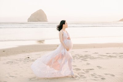 expecting mother portrait in the wind at the oregon coast. she in wearing a white and tan dress and has her hands on her belly
