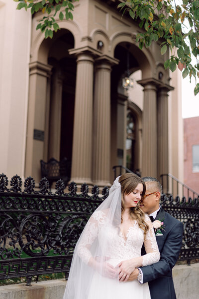 Romantic wedding portrait of the groom holding the bride from behind, with her veil draped over her shoulder, in front of Common House in Richmond, VA.