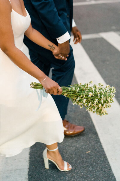 bouquet behind held by a bride in one hand as she holds her groom's hand with the other and walks across a crosswalk