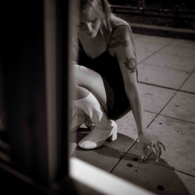 Black and white photo of a woman in a short dress and white boots crouching on a city sidewalk at night, partially obscured by a doorway, her tattooed arm extended as she reaches toward the ground with a contemplative expression.