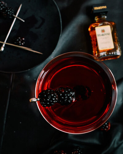 A moody and sophisticated image of a deep red cocktail garnished with fresh blackberries, set against a dark backdrop. Captured by Mica McCook, this photograph highlights the rich color and elegant presentation of the drink, creating a striking visual impact.