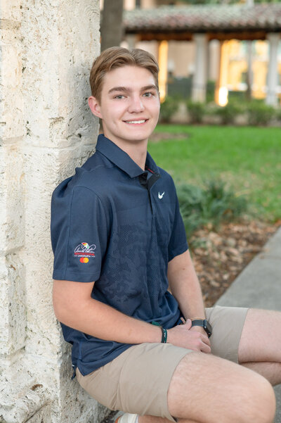 High school senior boy in blue polo shirt sitting against a pillar.