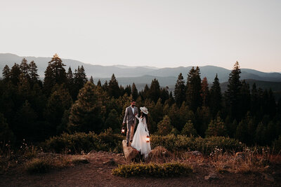 a bride and groom sitting on the ground, having a picnic with a mountain/forest view