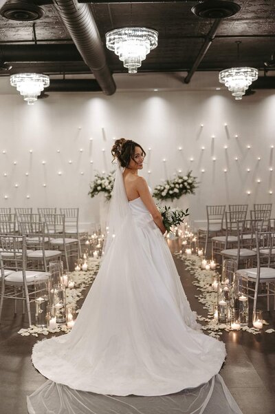 A bride in a strapless gown stands at the top of a petal-lined aisle at SoHo63, a Chandler wedding and event venue. Surrounded by candles, silver chairs, and a modern wall of hanging lights, she looks over her shoulder with a soft smile before the ceremony begins.