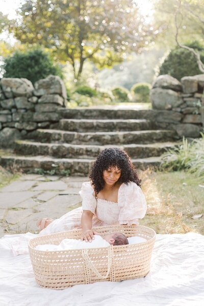 A mother in a pink toile dress sits next to her baby who is sleeping in a moses basket.