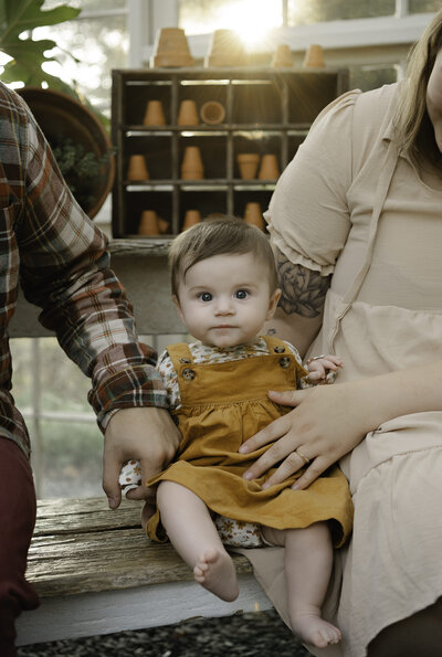 6 month old baby girl sits between her mother and father on a bench in the greenhouse at Prospect Farms in Lawrenceville Georgia