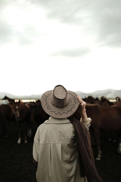 A person wearing a wide-brimmed hat and light coat stands facing a group of horses under a cloudy sky with mountains in the background, capturing the spirit of Sweet Pea Ink Creative’s unique brand design.