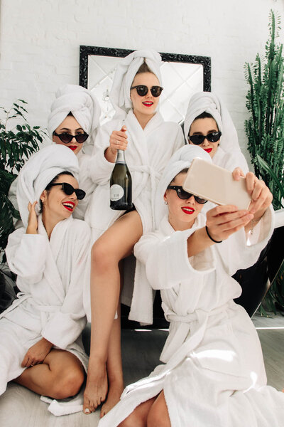 Group of women in white robes and towels taking a selfie while celebrating with champagne.