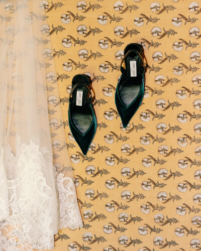 A fine art wedding flat lay featuring emerald green bridal shoes and a delicate lace veil, styled on vintage-patterned wallpaper at a luxury Austin wedding venue.