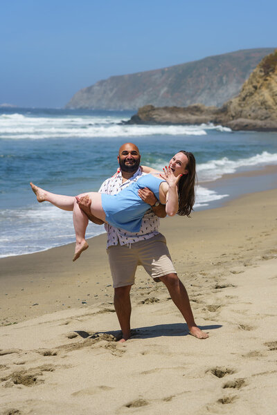 Man lifting his fiancée on the beach during an engagement session at Point Reyes, with ocean waves and coastal cliffs in the background