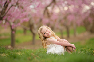 Little girl sitting in a blooming orchard smiling sweetly at the camera during photography session with Tiffany Hix in Boise
