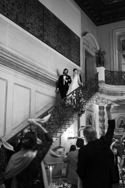 Couple descending the ornate staircase at Larz Anderson House in Washington, DC, to enter their wedding reception, with guests below cheering and waving napkins in celebration.