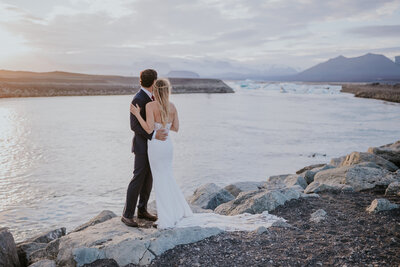 Romantic moment as newlyweds embrace on Iceland's glacier coast during sunset, surrounded by still waters and mountain silhouettes.