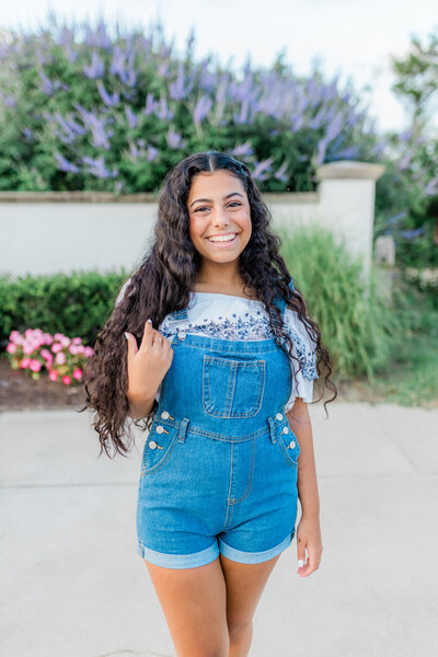 High school senior posing in front of flowers in Ocean City, New Jersey. She is wearing overalls and a mediterranean style shirt. Her hair is super long and curly and she is laughing.