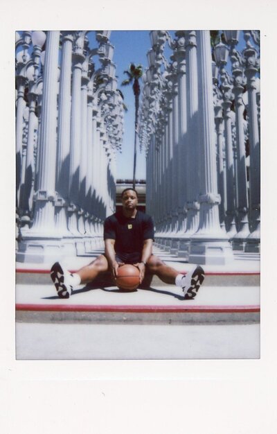 Jordan Foster sits on the steps of LACMA's famous outdoor light post installation while holding a basketball. Photographed by Renata Voci in bright sun lighting on Instax instant film.