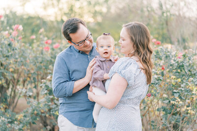 family photo of mom and dad wearing blue holding little girl in purple in a rose garden