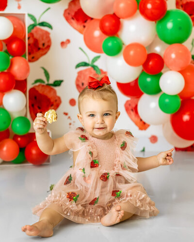 A little girl in a pink and red strawberry romper is holding a spoon in front of a strawberry-themed backdrop
