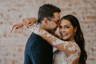 Brad and groom stand stomach to stomach as bride wraps her arms gently around grooms neck while smiling directly into the camera as her groom smiles at her