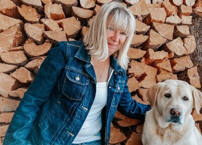 Susan Wyatt sitting with her Labrador, Flora, in front of stacked firewood, representing her expertise in landscape design and plant propagation.