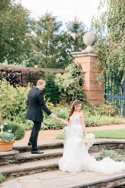 Bride and groom stroll hand-in-hand through a beautifully manicured garden, with the groom guiding the bride up stone steps. The bride wears a flowing, ruffled lace gown and holds a white floral bouquet, while the groom is dressed in a classic black suit. Surrounded by lush greenery, terracotta planters, and a brick wall, the scene exudes elegance and old-world romance.