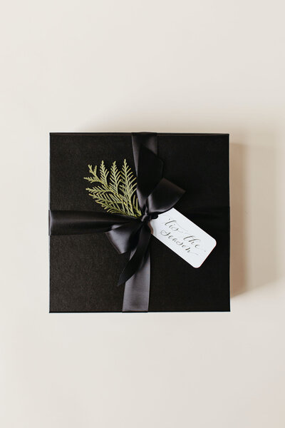 Black holiday gift box with black satin ribbon, sprig of greenery and custom calligraphy gift tag