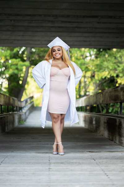 Senior girl wearing white cap & gown posing casually on a bridge