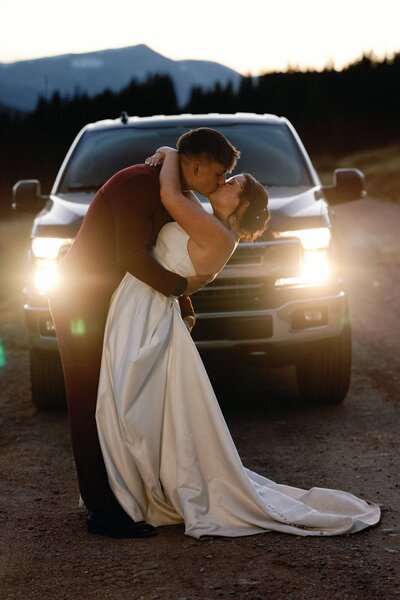 Bride and groom kiss in front of car headlights during mountain elopement.