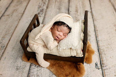 A peaceful newborn sleeps soundly on a tiny wooden bed, dressed in a soft cream-colored knit outfit with a matching hat. The cozy setup adds warmth and charm, making the scene both adorable and serene.