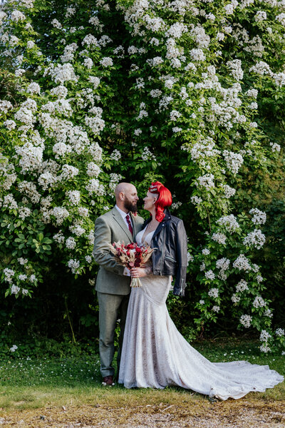Pin-up style bride and groom stand infont of a huge white floral bush at Coombe Manor Barns