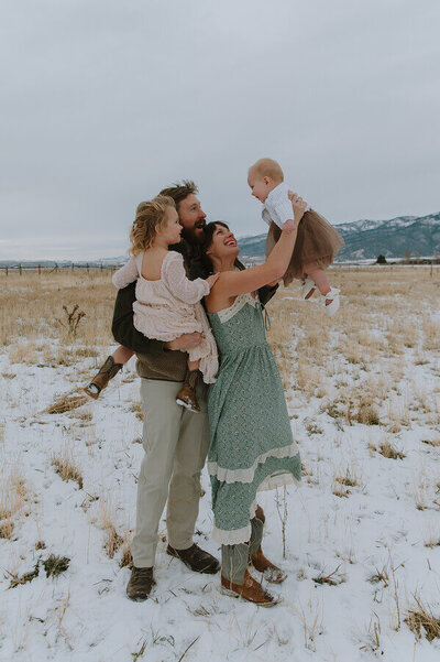 A mother holds her baby above her head smiling. Her husband is behind her holding their other daughter. They're all happy and enjoying the moment.