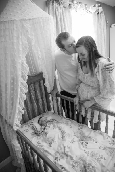 Father kissing wife's head while looking over crib | Taken by Newborn Maine Photographer