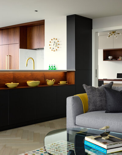 A living room space featuring a soft grey couch, glass coffee table, and black and oak cabinetry with an open shelf containing several decorative art pieces.
