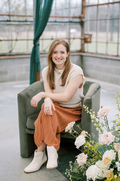 Dania, a wedding content creator with long hair sits smiling on a green armchair, wearing a cream top and rust skirt, surrounded by soft pink and white flowers indoors.