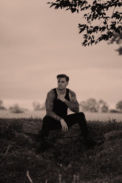 Tattooed guitar play sits in a field in a park in all black posing for an album cover.