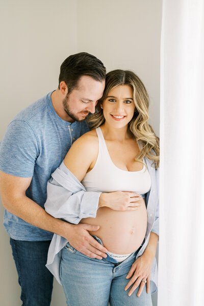 Expecting mom smiles at camera while husband holds her belly in maternity photo session